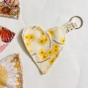 May include: A heart-shaped fabric keyring with a yellow floral pattern. The keyring has a cream background with small yellow flowers and brown stems. The heart is attached to a silver metal ring with a cream fabric loop.