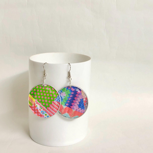 May include: A pair of round earrings with a silver metal frame and colourful patterned paper inside. The earrings are hanging from a white surface.