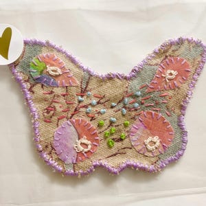 May include: A handmade butterfly-shaped fabric applique with a purple outline and a variety of colourful fabric scraps sewn together. The applique is on a beige background.