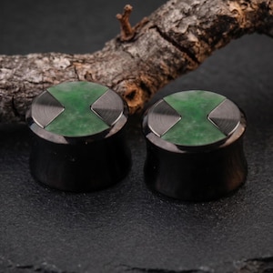 May include: A pair of circular ear plugs with a black metallic finish. The front face of each plug features a green stone inlay divided by two opposing brushed dark metal triangular segments, creating a distinct geometric pattern on the surface.