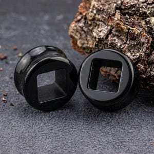 Puede incluir: Un par de dilatadores de oreja negros con un acabado liso y pulido. Cada pieza presenta un borde exterior circular y una abertura central cuadrada, diseñada como joyería de modificación corporal. Son de color negro sólido con brillo.