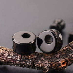 May include: Two smooth, glossy black ear plugs with a concave, saddle-shaped profile and a central circular hole. These circular body jewelry pieces feature a polished, reflective surface and a dark, solid color, designed for stretched ear piercings.
