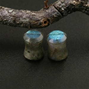 May include: Two labradorite stone ear plugs. Each plug has a rounded top with blue and green iridescent flashes. The plugs are a light gray color with darker gray and brown inclusions. The plugs are displayed on a black surface.