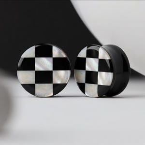 May include: Two black and white checkered ear gauges. The front of each gauge has a checkerboard pattern of black and white squares. The gauges are round and have a glossy finish. The background is black and white.