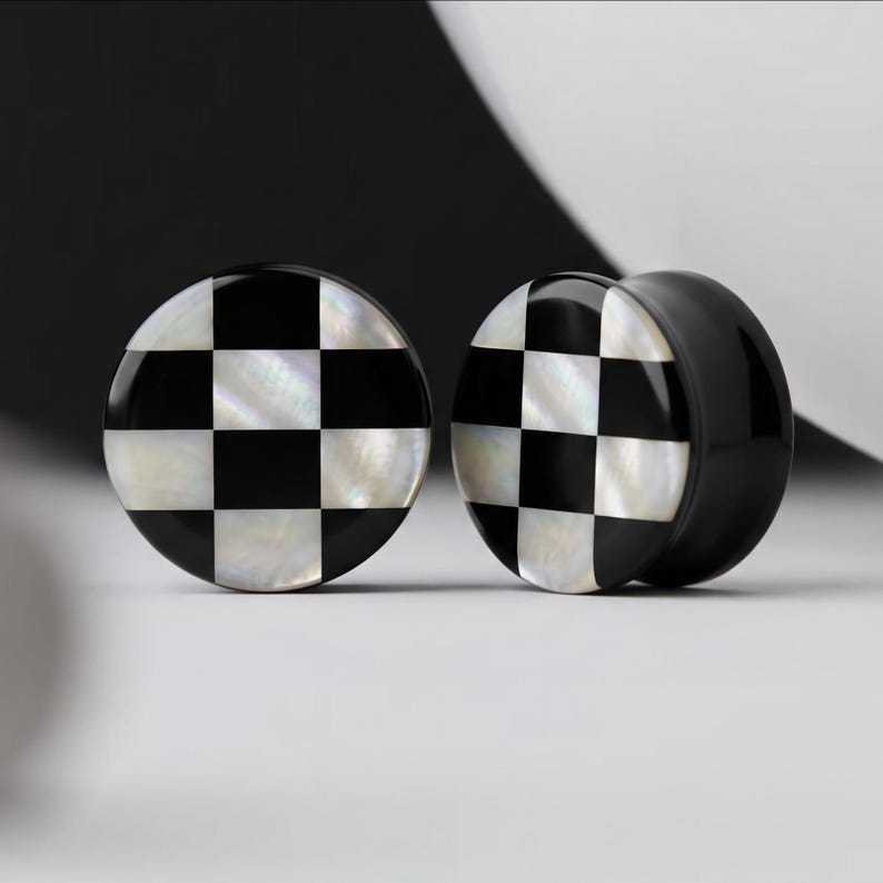 May include: Two black and white checkered ear gauges. The front of each gauge has a checkerboard pattern of black and white squares. The gauges are round and have a glossy finish. The background is black and white.