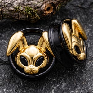 Peut inclure: Une paire d'écarteurs d'oreilles circulaires présentant un motif de tête de lapin doré poli avec des yeux noirs incrustés. Les têtes de lapin sont montées sur des tunnels cylindriques noirs lisses et brillants, offrant un contraste marqué.