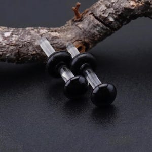 Puede incluir: Dos expansores de oreja negros con tallos transparentes y extremos redondeados. Los expansores se muestran sobre una superficie oscura con un trozo de madera en el fondo. Los expansores son de color negro brillante.
