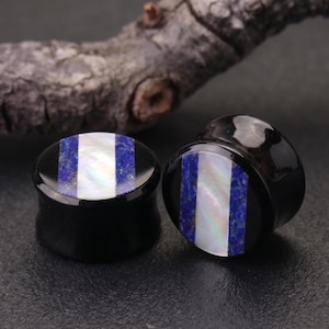 May include: Two black ear plugs with a striped design. The stripes alternate between white, blue, and black. The plugs are made of a glossy material and are displayed on a dark surface.