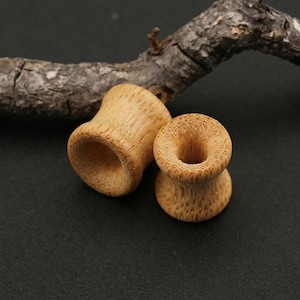 May include: Two light brown bamboo ear gauges. The gauges have a flared design with a cylindrical center. The surface of the gauges has a textured, dotted pattern. A dark brown tree branch is in the background.