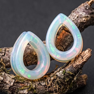 May include: A pair of teardrop-shaped ear plugs crafted from a translucent, milky white material with iridescent flashes of green, pink, and blue light throughout the surface, showcasing a smooth, polished finish and a hollow center design.
