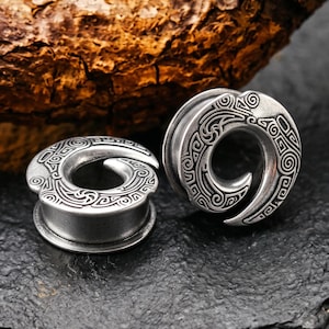 Op de afbeelding: Een paar zilverkleurige oorplugs met een cirkelvormig, op spiralen geïnspireerd ontwerp met ingewikkelde, gegraveerde patronen. Het sieraad heeft een gladde, metalen afwerking en een hol midden met een gedetailleerd, wervelend motief.