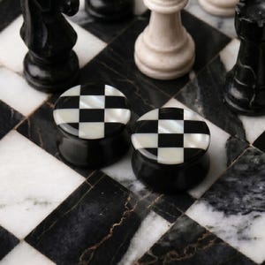 May include: Two black and white checkered plugs sit on a black and white marble chess board. The plugs have a black base with a checkered pattern of black and white squares on top. Chess pieces are in the background.