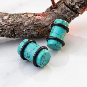 Puede incluir: Dos tapones para los oídos de piedra turquesa con anillos de goma negros. Los tapones tienen un aspecto veteado con vetas de color gris oscuro. Los tapones tienen forma cilíndrica y se muestran sobre una superficie blanca con un trozo de madera en el fondo.