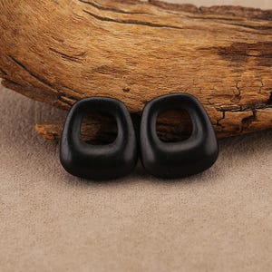May include: Two black, teardrop-shaped wooden earrings with a square hole in the center. The earrings are displayed on a piece of driftwood against a neutral background. The earrings have a smooth, polished finish.