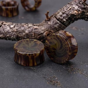 May include: A set of four circular ear plugs featuring a glossy, marbled pattern in shades of dark brown, amber, and tan. The ear plugs have a smooth, concave profile with flared edges, displaying a swirling, organic design throughout the surface.
