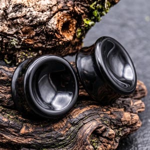 May include: A pair of circular black ear plugs with a smooth, concave surface and a polished finish. The plugs feature a flared edge design, creating a distinct silhouette. The dark, reflective material contrasts with the textured, organic surface.