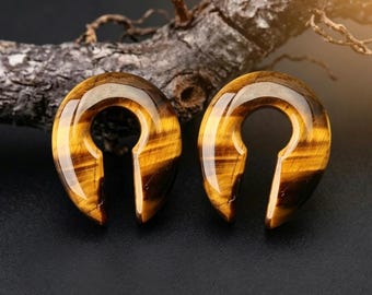 Puede incluir: Un par de dilatadores de oreja en forma de herradura hechos de piedra ojo de tigre pulida. Cada pieza presenta un acabado liso y brillante con bandas chatoyantes de color marrón dorado que recorren horizontalmente la superficie curva.