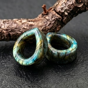 Puede incluir: Un par de dilatadores de oreja en forma de lágrima hechos de piedra labradorita pulida. El material muestra destellos iridiscentes de azul, verde y dorado en las superficies lisas y redondeadas, resaltando la estructura mineral natural.