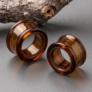 Puede incluir: Un par de dilatadores de oreja color ámbar con un acabado liso y translúcido y un diseño acampanado. Estas piezas de joyería corporal circulares presentan un tono cálido marrón anaranjado y una superficie pulida con un centro hueco.