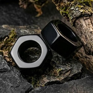 Puede incluir: Dos anillos negros lisos y pulidos con una forma exterior hexagonal y una abertura interior circular. Los anillos presentan una superficie brillante y reflectante, con un color negro sólido y uniforme en toda su estructura física.