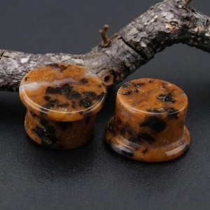 May include: Two orange and black stone ear plugs. The plugs have a cylindrical shape with a flared base. The stone has a mottled pattern of dark brown and orange hues. The plugs are displayed on a dark surface.