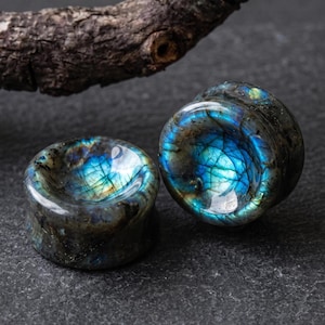 May include: A pair of circular ear plugs crafted from polished labradorite stone, featuring a concave center and a dark gray base color with iridescent blue and gold flashes visible across the smooth, reflective surfaces of the stone material.