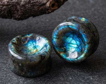 Op de afbeelding: Een paar ronde oorplugs gemaakt van gepolijste labradorietsteen, met een concaaf midden en een donkergrijze basiskleur met iriserende blauwe en gouden flitsen die zichtbaar zijn over de gladde, reflecterende oppervlakken van het steenmateriaal.