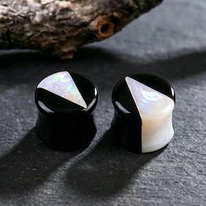 May include: A pair of circular ear plugs featuring a glossy black finish with a triangular inlay of iridescent white mother of pearl. The plugs have a smooth, concave profile and a polished surface that reflects light across the dark material.