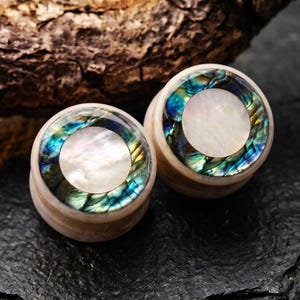 May include: A pair of circular ear plugs featuring a central white mother-of-pearl inlay surrounded by a ring of iridescent abalone shell with vibrant blue and green tones, set within a smooth, light-colored organic material frame.