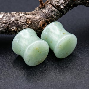 Op de afbeelding: Een paar gladde, lichtgroene stenen oorplugs met een uitlopend zadelontwerp. De plugs hebben subtiele natuurlijke minerale insluitingen en een gepolijste afwerking, met een lichte mintkleur en vage geelbruine spikkels over het oppervlak.