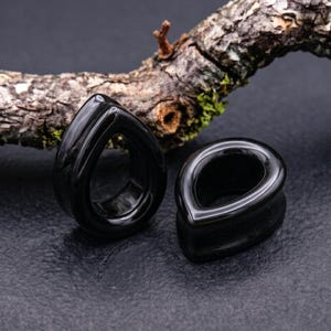 Puede incluir: Un par de dilatadores de oreja negros con un acabado liso y pulido en forma de lágrima. Estas piezas de joyería presentan un centro hueco y una superficie brillante que refleja la luz, mostrando un color oscuro sólido en toda la estructura.