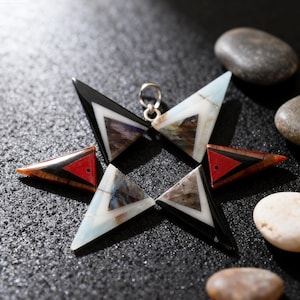 May include: A star-shaped pendant with six triangular points. Each point features a different design, including black, white, red, and patterned stone inlays. A silver loop is at the center for hanging. The pendant is displayed on a dark, textured surface.
