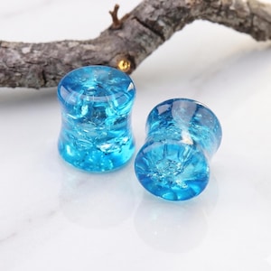 May include: Two clear blue glass ear plugs with a cracked ice effect. The plugs are cylindrical with a flared shape. The background is a white surface with a wooden branch.