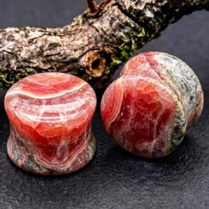 May include: A pair of circular ear plugs crafted from polished stone, featuring vibrant swirls of deep red, translucent pink, and white banding, with mottled grey and green mineral inclusions visible on the sides and edges.