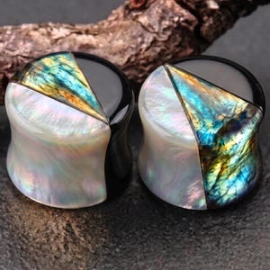 Puede incluir: Un par de expansores de oreja circulares con diseño dividido. Cada pieza combina una sección negra lisa, un segmento de concha blanca iridiscente y una incrustación triangular de labradorita azul y dorada con acabado pulido.