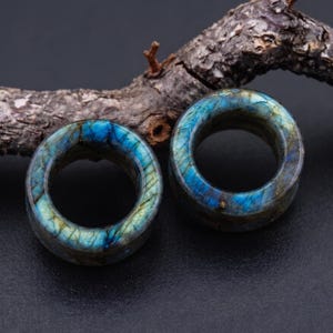 May include: A pair of circular ear plugs crafted from labradorite stone, featuring a polished surface with iridescent blue and gold flashes throughout the mineral structure, showcasing natural veining and a smooth, rounded cylindrical shape.
