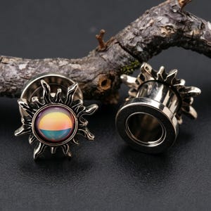 May include: Two silver-colored ear gauges with a sunburst design. The center of each gauge features a colorful, iridescent, circular inlay with shades of yellow, orange, and blue. The gauges are displayed on a dark surface with a piece of wood in the background.