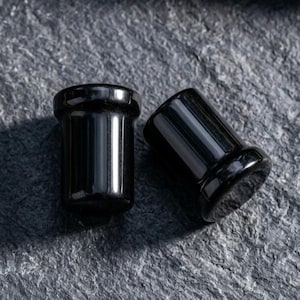 May include: A pair of solid black ear plugs with a smooth, glossy finish. Each plug features a cylindrical body with a slightly flared rim at one end, designed for body modification jewelry. The surface reflects light, highlighting the deep black color.