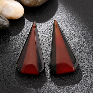 May include: Two geometric earrings with a triangular shape. Each earring features a combination of black and red-brown colors. The earrings have a hole at the top for hanging. The earrings are displayed on a dark, textured surface.