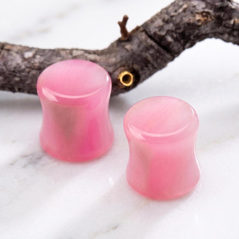 Pode incluir: Dois tamp&otilde;es de ouvido rosa polidos com um design alargado. Os tamp&otilde;es s&atilde;o de uma cor rosa suave e parecem ser feitos de um material liso e transl&uacute;cido. O fundo inclui uma superf&iacute;cie de m&aacute;rmore e um galho escuro e texturizado.