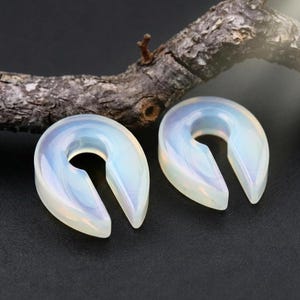 May include: Two iridescent, horseshoe-shaped ear gauges. The gauges are a translucent white with hints of blue and yellow, and are set against a dark background. A piece of wood is in the background.