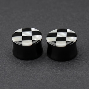 May include: Two black and white checkered ear plugs. The plugs have a black base with a checkered pattern of black and white squares on the top. The plugs are made of a glossy material.