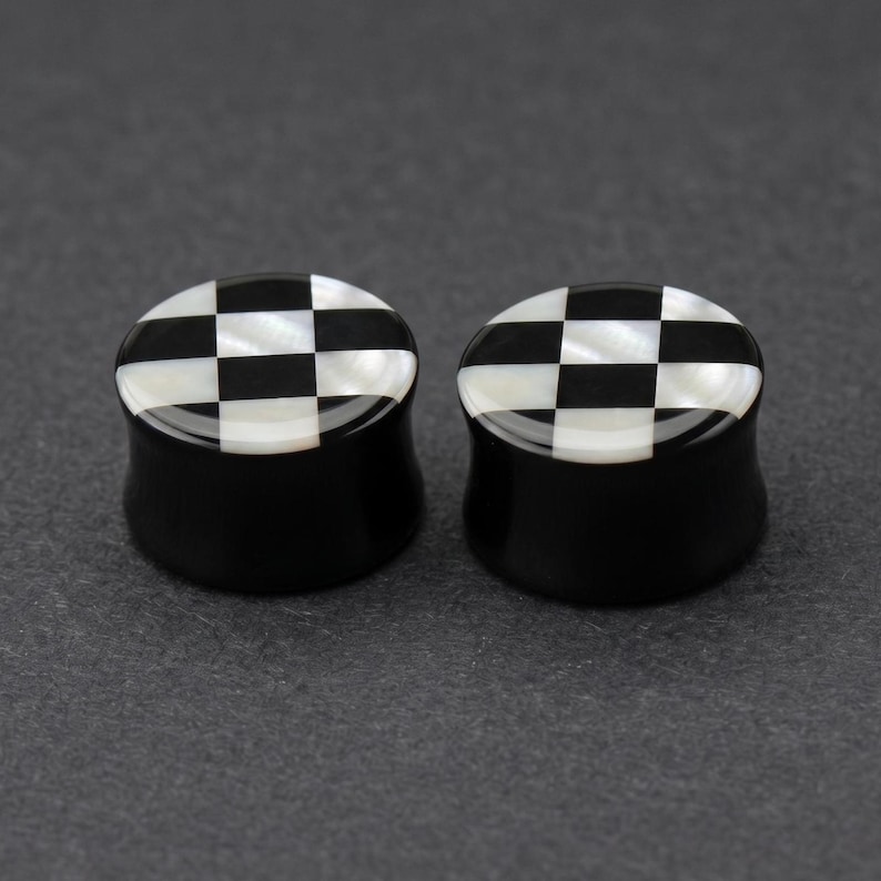 May include: Two black and white checkered ear plugs. The plugs have a black base with a checkered pattern of black and white squares on the top. The plugs are made of a glossy material.
