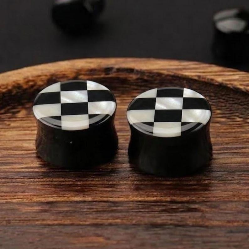 May include: Two black and white checkered plugs. The plugs have a black base with a checkered pattern of black and white squares on top. The plugs are sitting on a wooden surface.