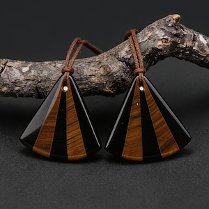 May include: Two triangular pendants with a striped pattern of black and brown tiger's eye. Each pendant has a small hole at the top with a brown cord. The pendants are displayed against a dark background and a piece of wood.
