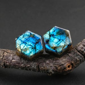 May include: Two hexagonal labradorite gemstones featuring a vibrant blue and teal iridescent flash with dark internal veining. The stones display a polished, smooth surface and are positioned side by side, showcasing their natural color variations.