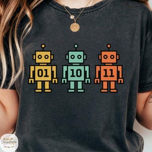 May include: Dark gray t-shirt featuring three colorful robot illustrations. Each robot is a different color: yellow, teal, and orange. The robots have the numbers 01, 10, and 11 printed on their chests.
