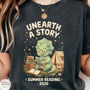 May include: Dark gray t-shirt featuring a cartoon dinosaur reading a book with the text "UNEARTH A STORY". The design includes a tent, books, and the words "SUMMER READING 2026". The dinosaur is green and wearing glasses.