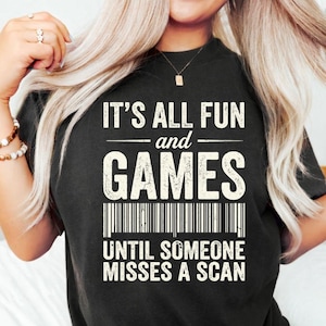 Può includere: T-shirt nera con la scritta bianca "IT'S ALL FUN and GAMES UNTIL SOMEONE MISSES A SCAN". La maglietta ha un design a codice a barre sotto le parole "GAMES". Girocollo.