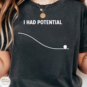 May include: Dark gray t-shirt with the white text "I HAD POTENTIAL" above a white line and a white ball graphic. The shirt is a casual style, suitable for everyday wear.
